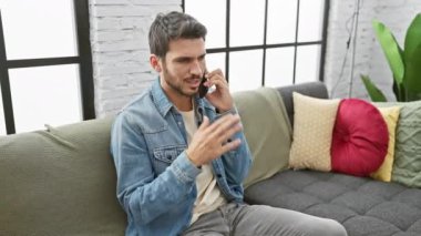 İspanyol bir adam telefonda, hareketli, sıkıntılı, kot giymiş, renkli bir koltukta oturuyor, içeride, gündüz vakti, tarafsız ifade, modern dekor..