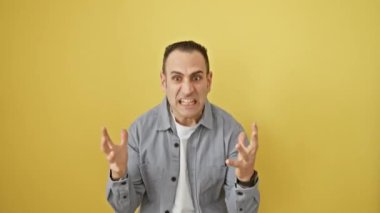 Sinirli İspanyol gömlekli genç adam öfkeyle bağırıyor, çılgın bir ifade takınıyor ve sarı izole edilmiş arka plana karşı kollarını kaldırıyor.