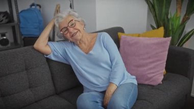 Gri saçlı ve gözlüklü gülümseyen son sınıf öğrencisi bir kadın, gri bir kanepede rahat rahat oturuyor..