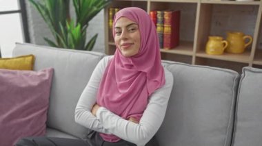 Sevinçli ve kendinden emin genç İspanyol kadın, neşe ve pozitiflik saçıyor, evdeki rahat bir kanepede dişlek bir gülümseme sergiliyor, güzel tesettürüyle Müslüman kimliğini kucaklıyor..