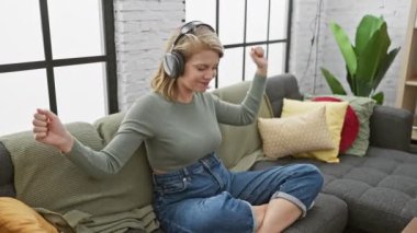 İçerideki kulaklıklarla müzik dinleyen, rahat bir oturma odasındaki kanepede uzanan genç bir kadın.