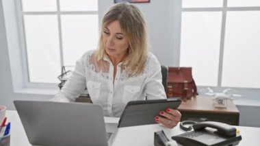 Modern ofis ortamında tablet ve laptop kullanan sarışın kadın iş, teknoloji ve profesyonellik taşıyor..