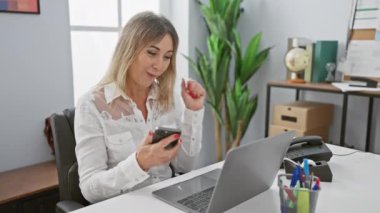 Neşeli, sarışın bir kadın, parlak, modern bir ofiste akıllı telefonunu kullanırken seviniyor..