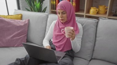 Başörtülü bir kadın dizüstü bilgisayarda çalışıyor ve modern oturma odasındaki kanepede kahve içiyor..