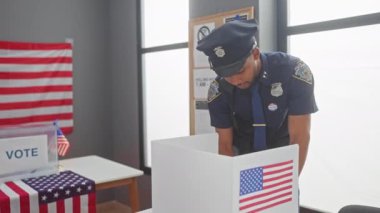 Afro-Amerikan polis memuru, seçim kulübesi ve bayrağı olan bir seçim merkezinde oy verirken gülümsüyor.