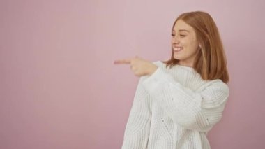 Rahat bir süveter giyen neşeli, genç, kızıl saçlı bir kadın, ayakta, pembe bir duvarda parmağını göstererek. Doğal, neşeli bir gülümseme ile, izole bir arkaplanda mutluluk getiriyor..