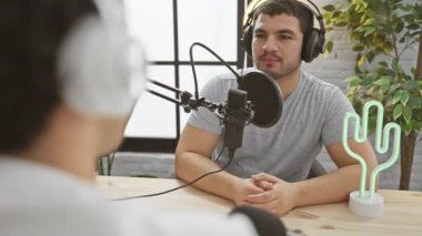 İki adam modern bir podcast stüdyosunda sohbet ediyordu. Biri mikrofona konuşuyordu..