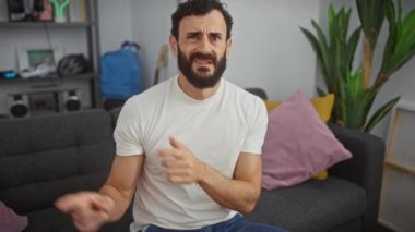Endişeli orta yaşlı bir adam evde, gergin bir şekilde kenara çekiliyor, beyaz bir tişört giyiyor, her iki eliyle şaşkınlığını ve endişesini dile getiriyor..