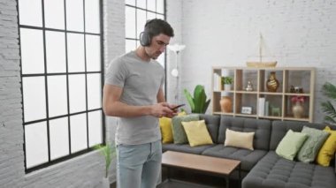 Genç bir İspanyol adam şık bir oturma odasında akıllı telefon kullanırken kulaklıkla müzik dinlemekten hoşlanıyor..