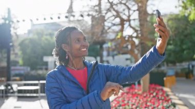 Gülümseyen Afrikalı bir kadın güneşli bir şehir ortamında selfie çekiyor, neşe ve şehir hayatı sergiliyor..
