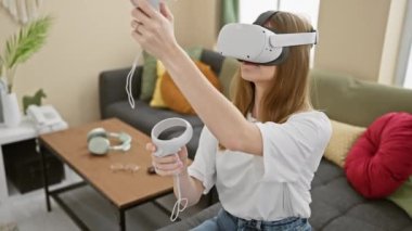 Modern bir oturma odasında, bir kulaklık ve kontrol cihazıyla sanal gerçekliği deneyimleyen genç bir kadın.