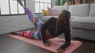 Renkli taytlı Afrikalı kadın rahat, modern bir oturma odasında yoga minderinde yan bacak kaldırma egzersizi yapıyor..