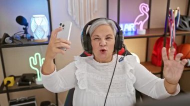 Kulaklık takan oyuncu, olgun bir kadın oyun odasında selfie çeker. Teknoloji ve son sınıf nişanı sergilenir..