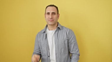 Tişört giyen neşeli, genç, İspanyol bir adam, üç parmak hareketini kendinden emin bir gülümsemeyle gösteriyor. İzole edilmiş sarı arka plana karşı pozitif bir kavram.