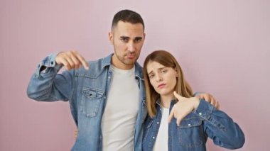 Bunalımlı kot pantolon ikilisi, kot gömlekli güzel bir çift pembe izole bir arka planda yan yana duruyorlar, üzüntülerini ifade ediyorlar ve parmaklarıyla aşağıdaki duyguları gösteriyorlar.