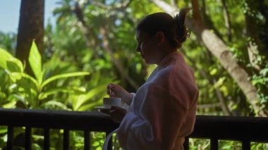 Huzurlu genç bir kadın güneşli bir terasta kahvenin tadını çıkarıyor. Etrafı yemyeşil bale ile çevrili..