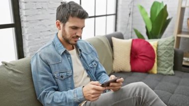 Gündelik kıyafetli genç bir İspanyol, modern bir oturma odasında rahat bir şekilde gülümseyen akıllı telefonunun tadını çıkarıyor..