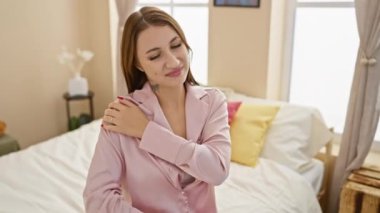 Pembe tişörtlü, suratını buruşturan genç bir kadın omzunu sımsıkı kavrıyor, iyi aydınlatılmış bir yatak odasında bir yatakta oturuyor..