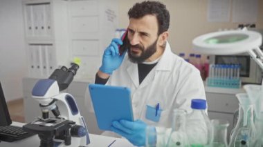 Laboratuvar önlüklü orta yaşlı sakallı bir adam. Laboratuvar ortamında tablet ve telefon kullanıyor..