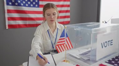 Gözlüklü beyaz bir kadın kapalı bir ABD seçim merkezinde anket görevlisi olarak görev yapıyor. Amerikan bayrağı arka planında..