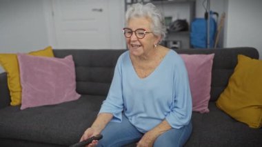 Kanepede renkli yastıklarla oturan gülümseyen yaşlı kadın oturma odasında uzaktan kumandayla televizyon tutuyor..