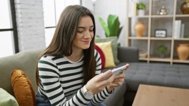 Gülümseyen genç bir kadın rahat, modern bir oturma odasında akıllı telefon kullanır ve günlük ev hayatını yakalar..