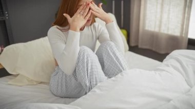 Genç, sıkıntılı kızıl saçlı bir kadın bir yatakta oturuyor, elleriyle yüzünü örtüyor, yatak odası düzeninde..