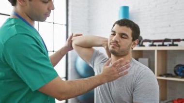 Klinikte bir erkek terapist, genç bir adama içeride omuz rehabilitasyonu egzersizinde rehberlik eder..