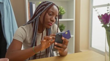 Saç örgülü Afrikalı kadın evde kredi kartı ve akıllı telefonuyla online alışveriş yapıyor..