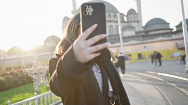 Gün batımında İstanbul caddesinde gözlüklü ve akıllı telefonlu gülümseyen genç kadın.