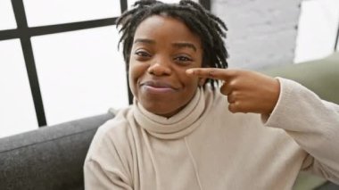 Büyüleyici genç siyahi kadın evdeki kanepede dinleniyor, parmağını yüzüne ve burnuna doğrultuyor, ikincil olarak güzellik ve cilt sağlığını gösteriyor. Işıl ışıl gülüşü odayı aydınlatıyor.