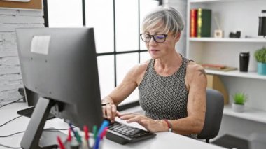 Modern ofis ortamında bilgisayar başında çalışan gri saçlı olgun bir kadın..
