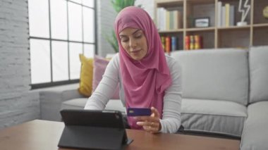 Sıcak oturma odasında online alışveriş için tesettürlü gülümseyen kadın kredi kartı ve tablet kullanıyor..