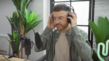 Modern bir stüdyoda sakallı yakışıklı bir adam mikrofona kulaklıkla gülümsüyor.