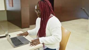 Ofis ortamında sırtına dokunan örgü ören Afrikalı Amerikalı bir kadın, bir dizüstü bilgisayarın yanında, ağrıyı işaret ediyor..