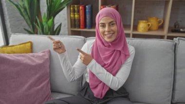 Neşeli, tesettürlü genç İspanyol kadın kendinden emin bir şekilde yan tarafı işaret ediyor, evdeki kanepesinde gülümsüyor, bizi mutlu bir şekilde oturma odasına davet ediyor, kameralara yakalanan samimi bir karşılama.