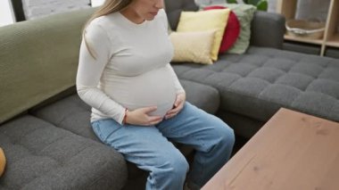 Genç bir İspanyol hamile kadın içeride oturuyor, karnına dokunuyor ve rahat bir oturma odasında düşünceli görünüyor..