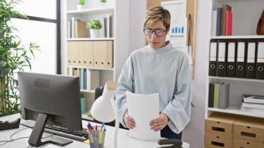 Kısa sarı saçlı, kendine güvenen İspanyol kadın modern ofis ortamında dokümanları gözden geçirir, profesyonellik yayar ve odaklanır..