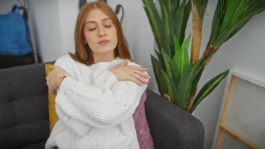 Neşeli, genç, bekâr, kızıl saçlı kadın, ev içinde, kendine güveni ve sevgisini ifade ediyor. rahat bir kazak giyiyor, mutlu bir gülümseme yayıyor, kendine sarılıyor. Kendi dairesinde mutluluğun portresi.