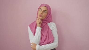 Düşüncelere dalmış, tesettürlü genç İspanyol kadın, parmaklar çeneye, gözler yukarı bakıyor, izole pembe arka planı merakla ve şüpheyle boyuyor.