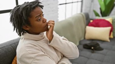 Gergin, siyah saçlı bir kadın, evdeki kanepede tırnaklarını yiyor. Stres ve anksiyete sorunu yaşıyor. 