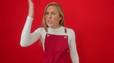 Öfkeli genç bir kadın, sarışın, kırmızı arka planda dikilmiş, öfkeyle bağırıyor, önlük giyiyor, sinirli, hayal kırıklığına uğramış, agresif.