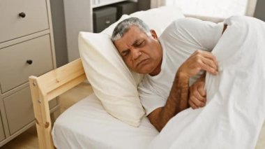 Orta yaşlı bir adam düzenli bir yatak odasında yatağın altında konfor buluyor, günlük ev hayatını resmediyor..