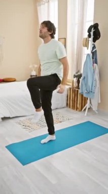 Sıradan giyinen genç sakallı bir adam, yatak odasındaki mavi bir paspasın üzerinde yoga yapıyor. Sağlığını ve evdeki refahını gösteriyor..