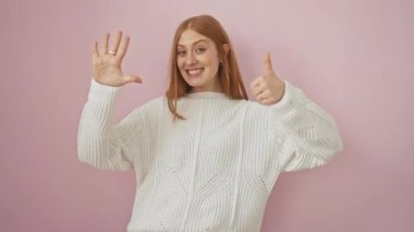 Neşeli, genç, kızıl saçlı kadın parmaklarıyla 6 'lık çakıyor. Gülümsüyor ve kendine güveniyor. Pembe bir arka planda izole edilmiş pembe bir süveter giyiyor.