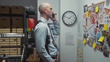 Sakallı, kel dedektif ofiste telefonda kanıt panosu ve saatle konuşuyor..