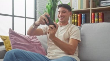 Gülümseyen İspanyol genç adam, renkli yastıklarla dolu bir oturma odasında gri bir kanepede rahatça otururken telefonuyla oyun oynamaktan zevk alıyor..