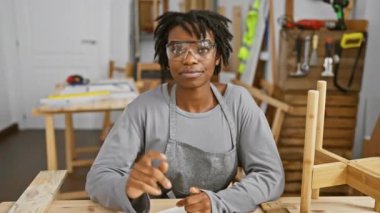 Rastalı neşeli siyah kadın, marangozluğa güveni tam, sallanan güvenlik gözlükleri, gülerken bir yandan da dostça bir jest yapıyor.