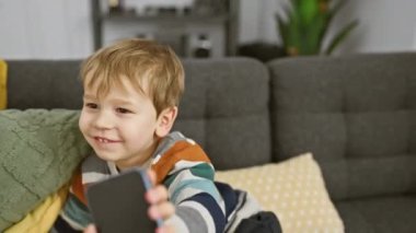 Hoş bir oturma odasındaki yastıklı gri koltukta otururken akıllı telefon tutan neşeli sarışın bir çocuk..