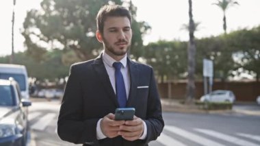 Sakallı iyi giyimli bir adam güneşli bir caddede akıllı telefon kullanıyor..